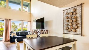 A dining area with a dark wooden table and white chairs, a TV mounted on the wall, and a decorative shell wall art piece. - Convenient condo with WiFi, W/D, shared pool, tennis, & bocce, and more (Panama City Beach)