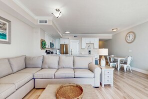 A modern living room with a sectional sofa, a dining area with a table and chairs, and a kitchen in the background. - Beachfront condo with hot tub, sauna, pools, and stunning ocean views (Panama City Beach)