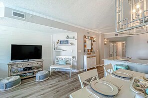 A living room with a television, white furniture, and a dining area with a table set for four. - Beautifully decorated oceanfront condo with resort pool, & huge walk-in showers (Santa Rosa Beach)