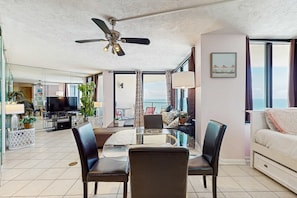 A modern living room with a glass dining table, two chairs, a ceiling fan, a TV, and a balcony with a view. - Snowbird-friendly condo in Panama City Beach with shared pool (Panama City Beach)