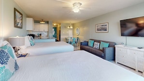 Quarto de hotel com duas camas, um sofá, uma TV e uma cozinha compacta.