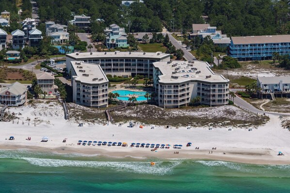 Un hotel frente a la playa con varias construcciones, piscina y una playa de arena con sombrillas.