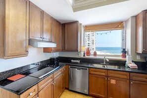 Eine Küche mit Holzschränken, einer schwarzen Arbeitsplatte und einem Geschirrspüler. Ein Fenster bietet Blick auf den Strand und ein Gebäude.
