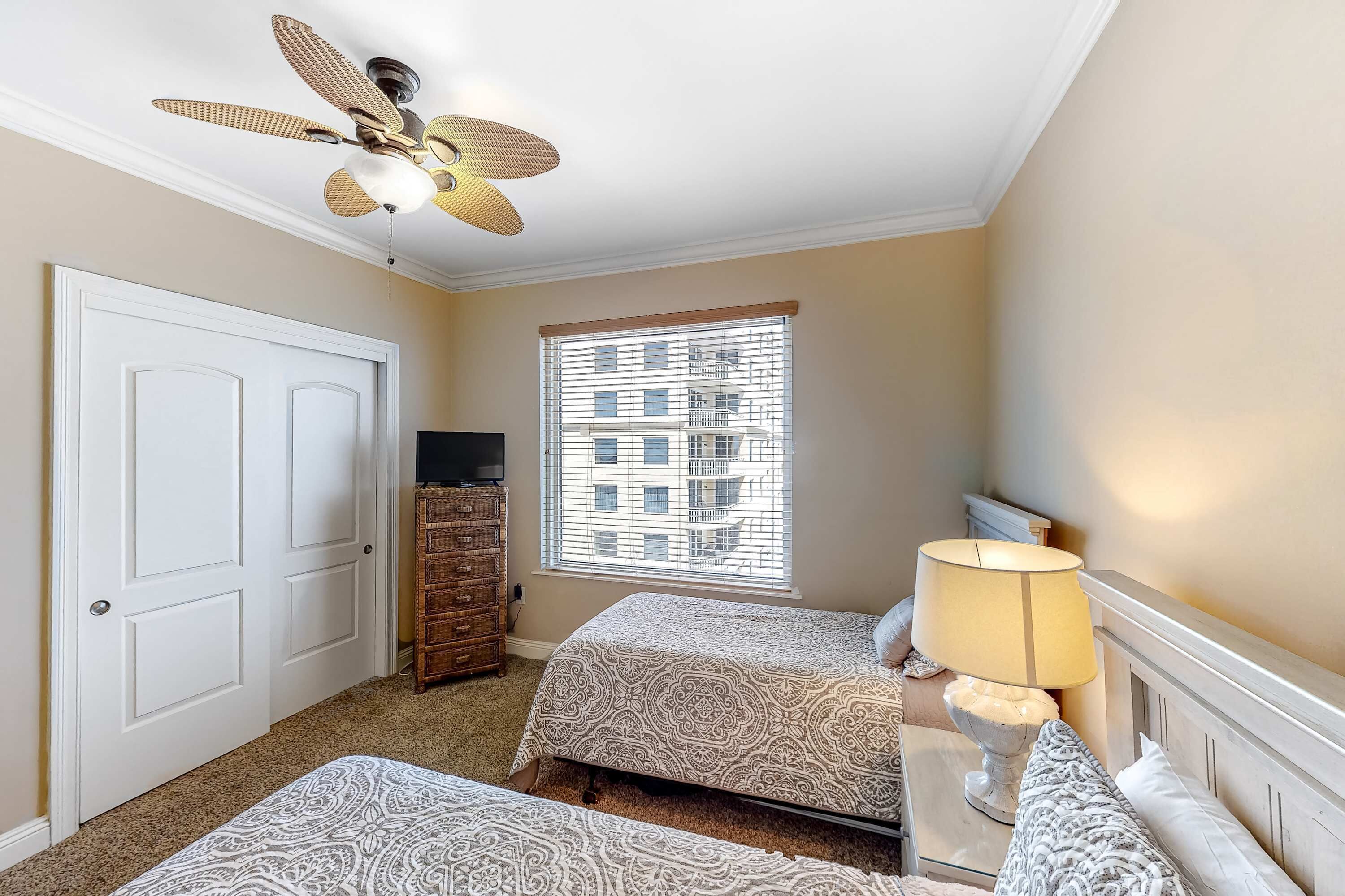 Un dormitorio con dos camas, ventilador de techo, ventana con persianas, cómoda y lámpara.