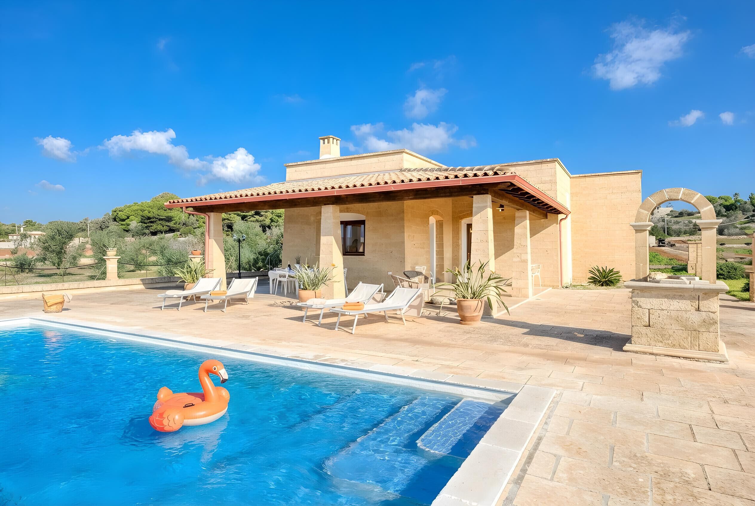 A pool with a flamingo float, lounge chairs, and a stone building with a tiled roof.