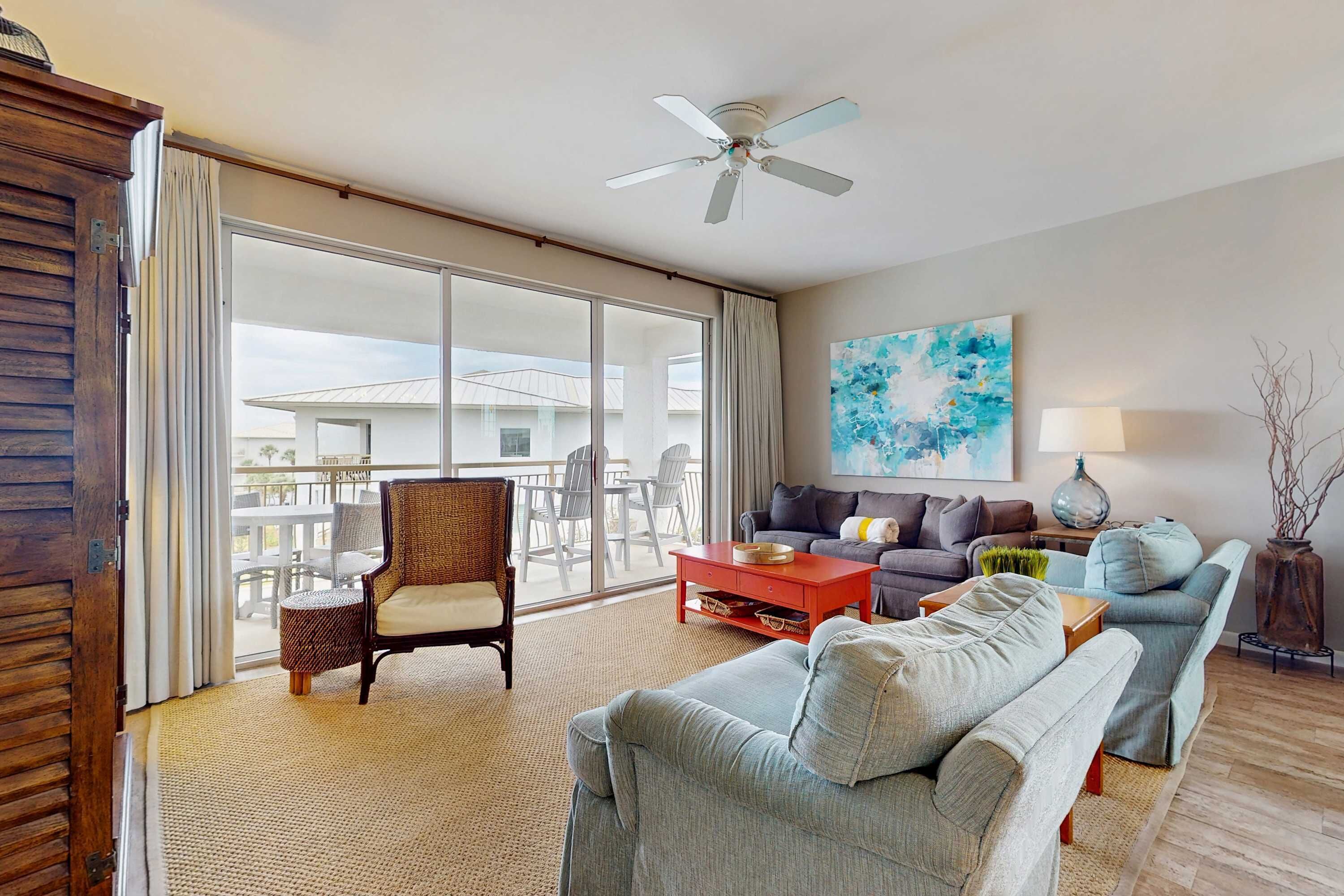 A spacious living room with a large sliding glass door leading to a balcony, a ceiling fan, a modern abstract painting, and a cozy seating area with a red coffee table.