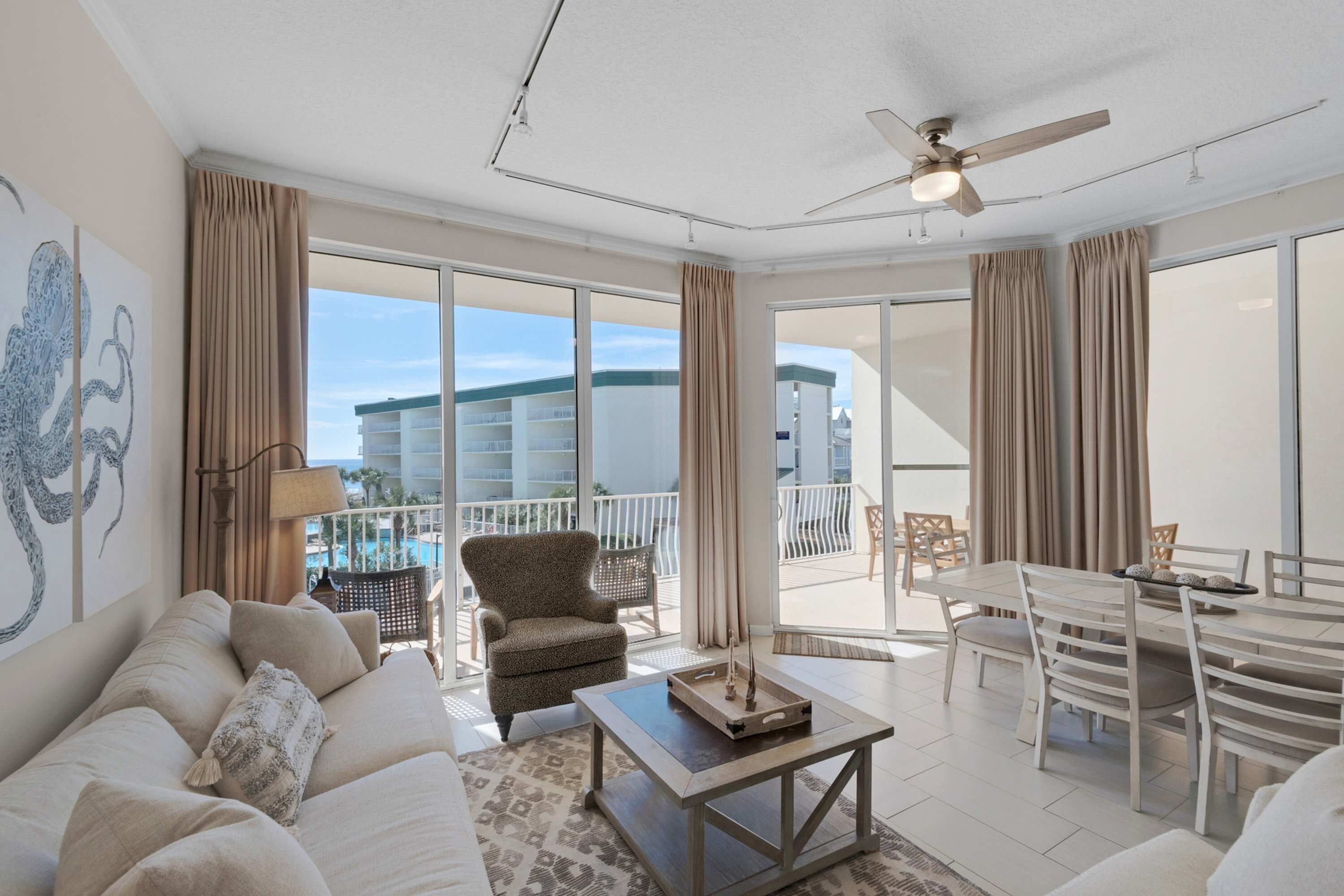 A spacious living room with a sofa, armchair, and dining table. Large windows provide a view of a building and a pool.