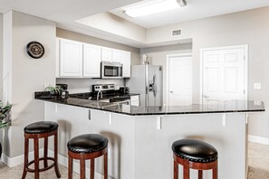 Una cocina con armarios blancos, encimera negra, refrigerador de acero inoxidable y dos taburetes de bar.