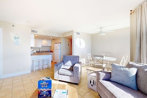 A modern living room with a grey sofa, a coffee table, and a dining area with a table and chairs. There is a kitchen in the background with white cabinets and a refrigerator. - Incredible beachfront getaway with views, pools, & tennis - Snowbirds welcome (Panama City Beach)