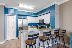 A modern kitchen with white cabinets, a stainless steel refrigerator, a microwave, and a granite countertop. There are four bar stools with wooden seats and black metal frames. - Picturesque gulf-front views & on-site pools, hot tubs, and beach service (Panama City Beach)