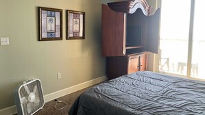 A bedroom with a bed, a standing fan, and two framed pictures on the wall. - Cozy beachfront condo with private balcony, shared pool & beach access (Panama City Beach)