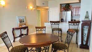 A dining area with a round wooden table and six chairs with patterned upholstery. - Cozy beachfront condo with private balcony, shared pool & beach access (Panama City Beach)