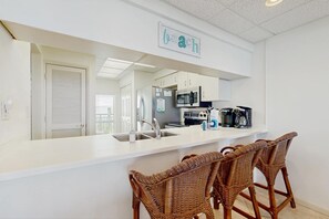 A modern kitchen with white cabinetry, a stainless steel sink, and a microwave. There are wicker chairs at the counter and a sign above the sink. - Gorgeous beach condo with furnished balcony, panoramic view & pickleball (Seacrest)