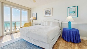 Habitación de hotel con una cama grande, ventilador de techo, vista a la playa y una mesa con lámpara.