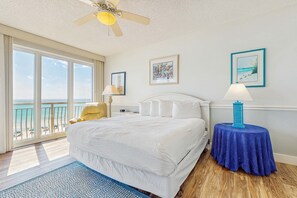 Habitación de hotel con una cama grande, ventilador de techo, vista a la playa y una mesa con lámpara.
