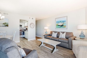 A modern living room with a grey sofa, a white coffee table, and a grey armchair. A kitchen is visible in the background. - Gulf-front condo with amazing views, WiFi, central AC, pools, & fitness room (Panama City Beach)