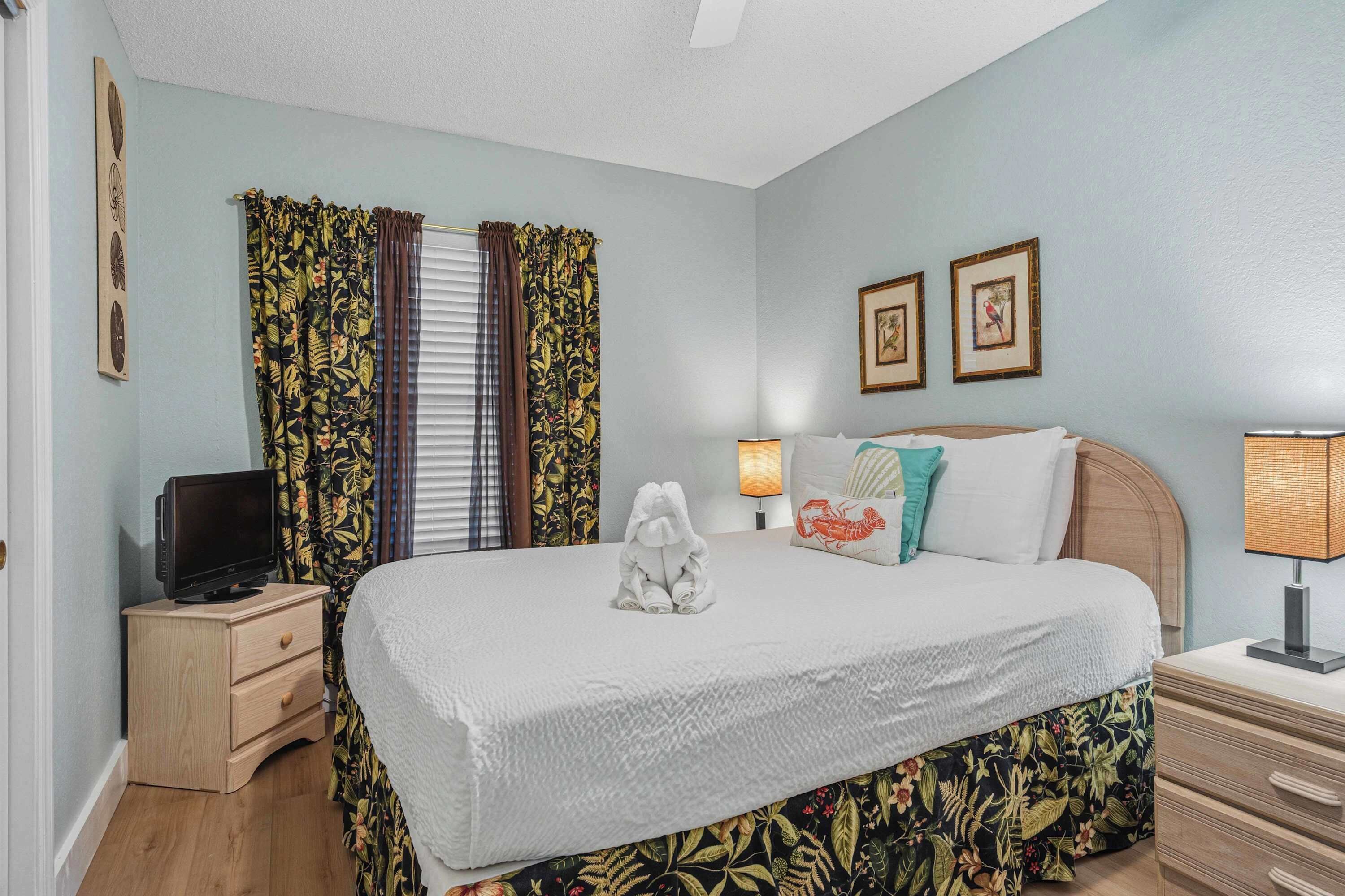 A bedroom with a bed, curtains, a television on a nightstand, a lamp, and two framed pictures on the wall.
