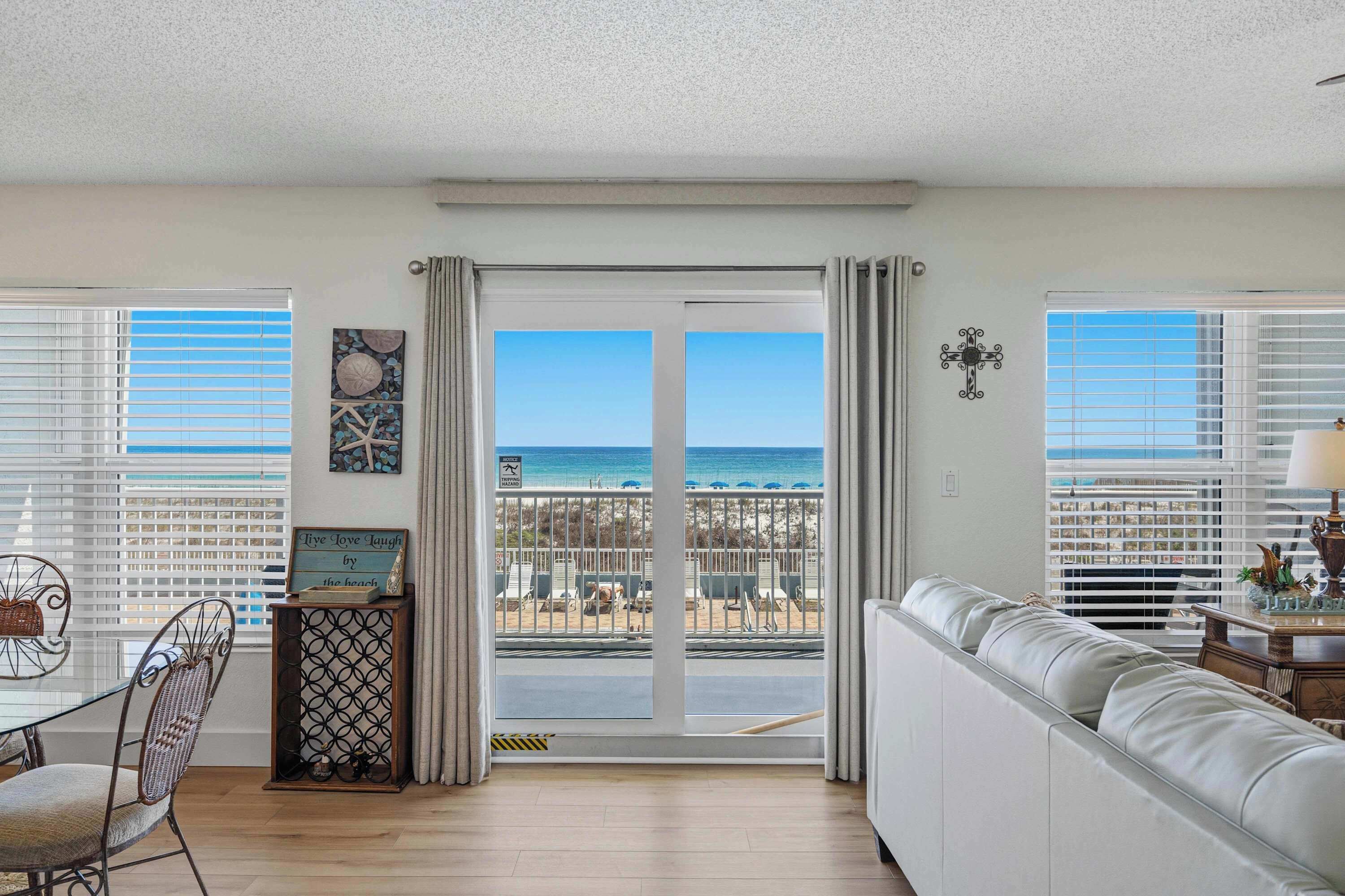 Ein Zimmer mit Blick auf den Strand, eine Schiebetür, ein weißes Sofa, ein Esstisch mit Stühlen und eine Wanddekoration.