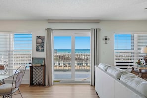 A room with a view of the beach, a sliding glass door, a white sofa, a dining table with chairs, and a decorative wall piece. - Spacious condo in with free WiFi, pool access, central AC, and washer&dryer (Fort Walton Beach)