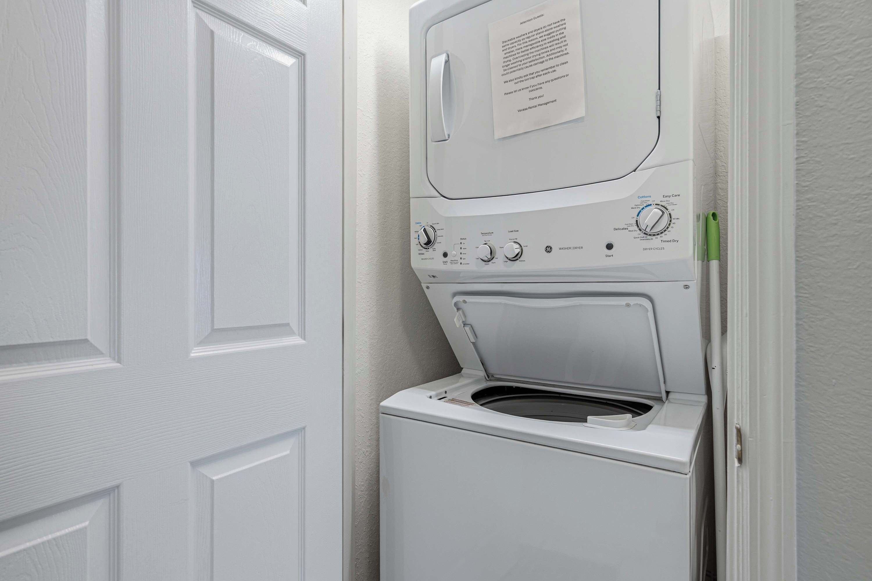 A front-loading washing machine in a laundry room with a door and a wall-mounted instruction manual.