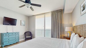Ett hotellrum med en stor säng, en takfläkt, en TV, en byrå, en stol, en balkong med gardiner och en lampa.