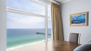 A room with a large window overlooking a beach and ocean, a wooden table, and a framed painting on the wall. - Beachfront resort condo with Gulf views, outdoor pool, & private balcony (Panama City Beach)
