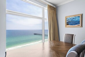 Ein Zimmer mit großem Fenster mit Blick auf Strand und Meer, einem Holztisch und einem gerahmten Gemälde an der Wand.