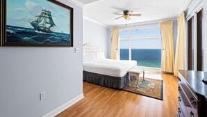 A bedroom with a bed, a ceiling fan, a large window with curtains, and a painting of a ship on the wall. - Beachfront resort condo with Gulf views, outdoor pool, & private balcony (Panama City Beach)