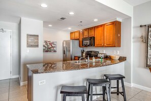 A kitchen with granite countertops, wooden cabinets, stainless steel appliances, and two bar stools. - Luxurious beachfront condo with ocean views, Pool & Hot tub (Panama City Beach)