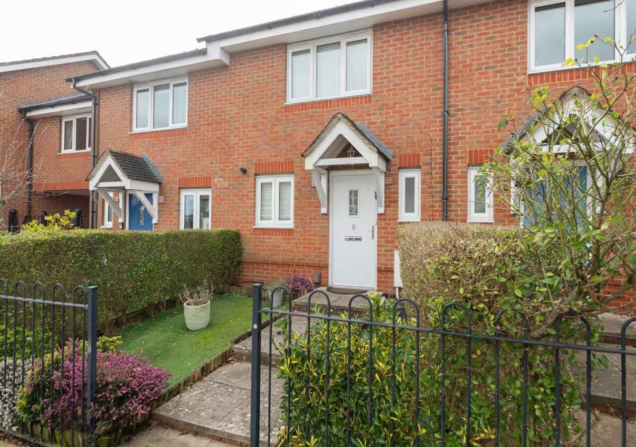 A red brick terraced house with a white front door, a small front garden, and a metal fence.