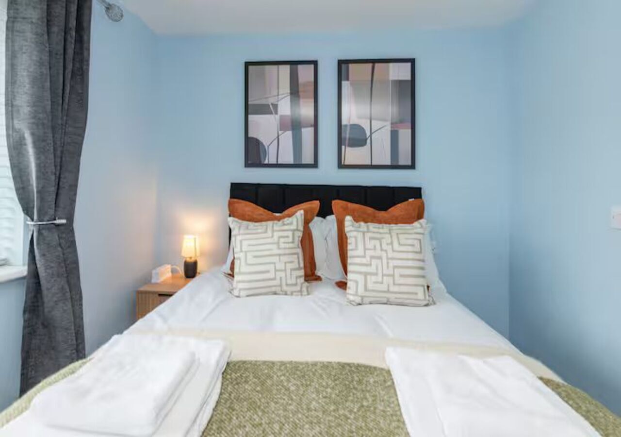 Una cama bien hecha con sábanas blancas y almohadas estampadas, una lámpara de noche y una obra de arte abstracta en la pared.
