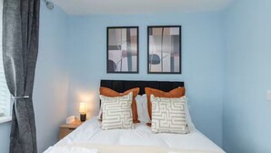 Una cama bien hecha con sábanas blancas y almohadas estampadas, una lámpara de noche y una obra de arte abstracta en la pared.