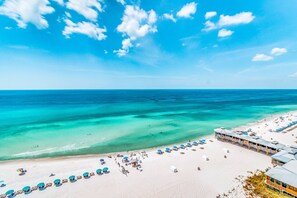 An aerial view of a beach with clear turquoise waters, a sandy shore, and a resort with multiple beach umbrellas and lounge chairs. - Beachfront condo with WiFi, balcony, central AC, pools, tennis, & fitness room (Panama City Beach)