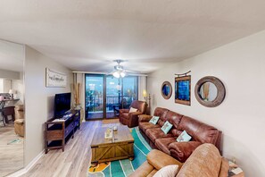 Un salón espacioso con un sofá de cuero marrón, una mesa de centro de madera y un televisor sobre un mueble. Se aprecia una ventana grande con vistas al exterior.