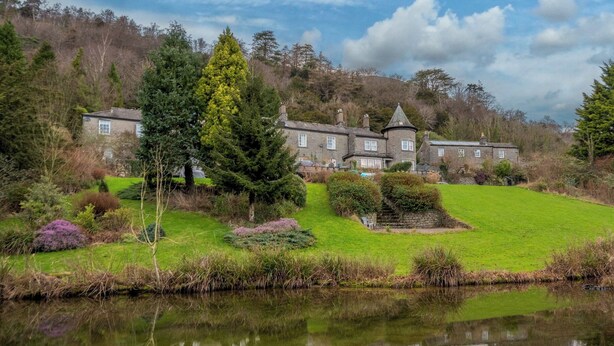 A large stone building with multiple windows, surrounded by lush greenery and a pond.