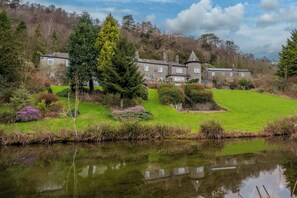 A large stone building with multiple windows, surrounded by lush greenery and a pond.