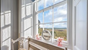 A room with a large window offering a view of a field and hills. A bench with a mirror and flowers is placed by the window.