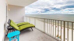 En balkong med två gröna fåtöljer och en blå stol, med utsikt över en strand med parasoller och en klar himmel.