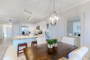 A modern kitchen with white cabinets, a stainless steel refrigerator, and a marble countertop. There is a wooden dining table with two white chairs and two potted plants. - Gorgeous condo with pool & Gulf view, stunning decor, bunk beds & pickleball (Santa Rosa Beach)