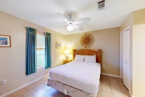 Une chambre à coucher comprenant un lit, un ventilateur de plafond, une fenêtre avec des rideaux, un élément de décoration murale et une table de chevet avec une lampe.
