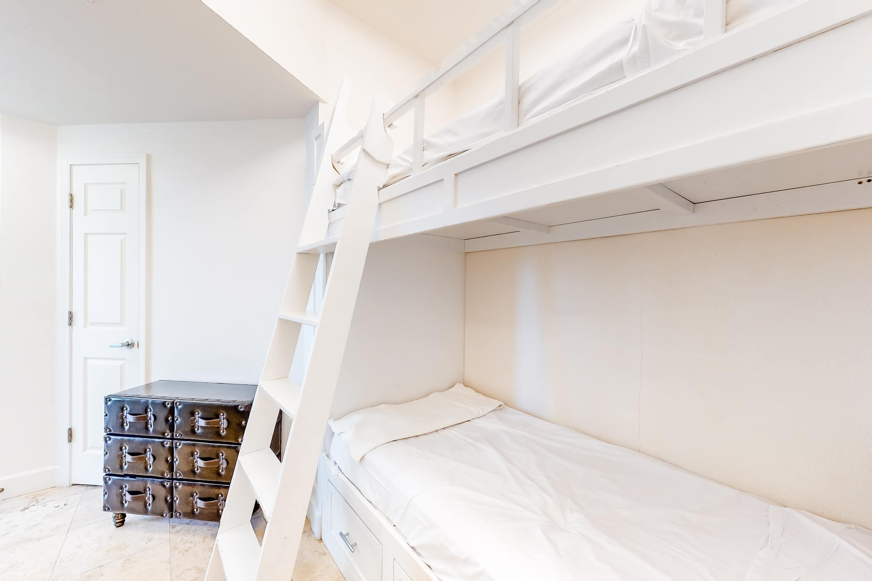 Un lit superposé avec une literie blanche, une porte blanche et une commode dotée de plusieurs compartiments.