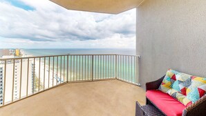 A balcony with a view of the beach and ocean, a wicker sofa with a colorful cushion, and a concrete floor. - Beautiful 28th-floor condo w/shared gym/Roman spa, hot tub, two shared pools (Panama City Beach)