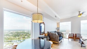 A spacious living area with a large round table and chairs, a sofa set, and a view of a cityscape through large windows. - Beautiful 28th-floor condo w/shared gym/Roman spa, hot tub, two shared pools (Panama City Beach)