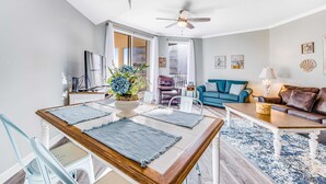 A modern living room with a dining area, featuring a ceiling fan, a blue sofa, a coffee table, and a dining table with placemats. - First-floor condo with sauna, gym, pool, tennis & pickleball (Santa Rosa Beach)