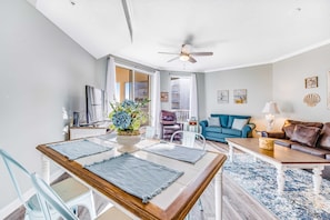 Ein modernes Wohnzimmer mit Essbereich, ausgestattet mit einem Deckenventilator, einem blauen Sofa, einem Couchtisch und einem Esstisch mit Platzdeckchen.