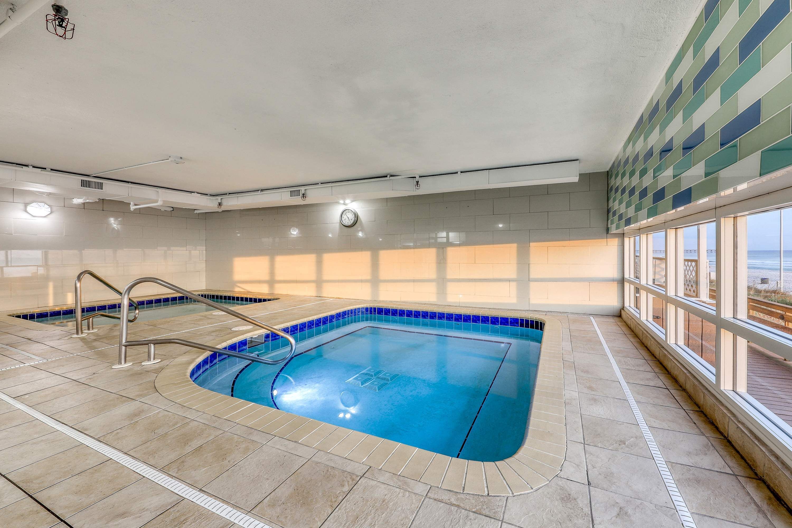 An indoor swimming pool with clear water, a metal ladder, and a view of the sea through large windows.