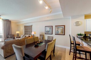 A modern living room with a dining table, sofas, and chairs. There is a kitchen area with a countertop and a clock on the wall. - Warm & welcoming condo w/Gulf and beach views, on-site pools & hot tub (Panama City Beach)