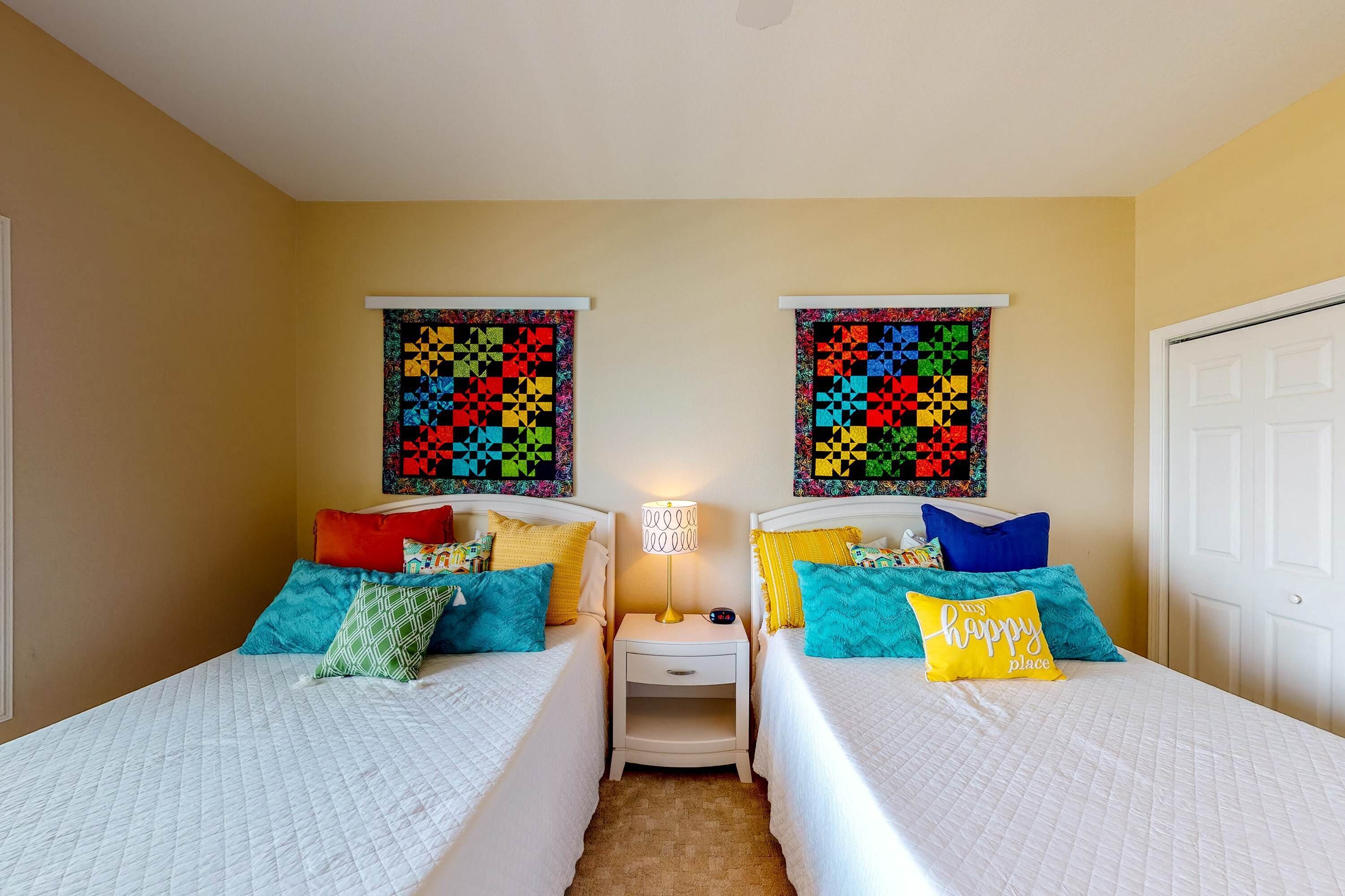 Una habitación con dos camas, cada una con ropa de cama y almohadas coloridas, una mesita de noche con lámpara, y dos colchas colgadas en la pared.