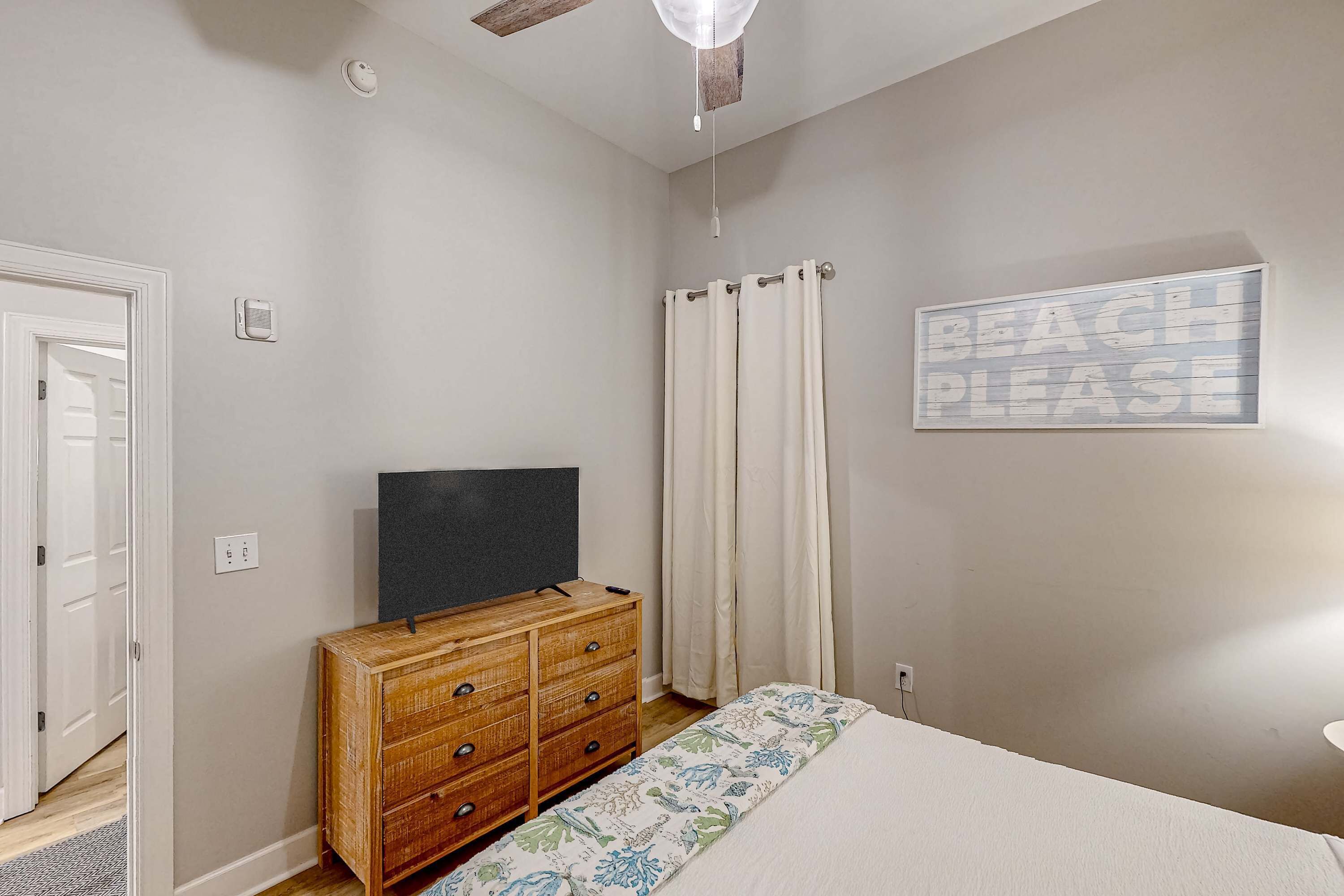 A bedroom with a bed, a wooden dresser, a television, a 'BEACH PLEASE' sign, and a door.