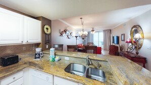 A kitchen with granite countertops, a double sink, and white cabinetry. There is a coffee maker, a toaster, and a dish rack on the counter. - Coastal condo steps to beach with pools, hot tub, gym & amazing views (Perdido Key)